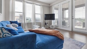 A modern living room with a blue sofa, a flat-screen TV, and large windows with blinds. - Monthly Stays Luxe Des Peres Suite with Complimentary Starbucks Onsite!  (Des Peres)