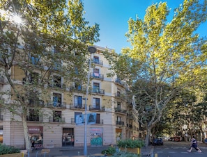 Un edificio de varios pisos con balcones, una calle arbolada y un cielo azul despejado.