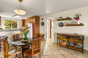 Une cuisine moderne avec des meubles en bois, une table à manger avec des chaises, un luminaire suspendu et une vue sur l’extérieur par une fenêtre.