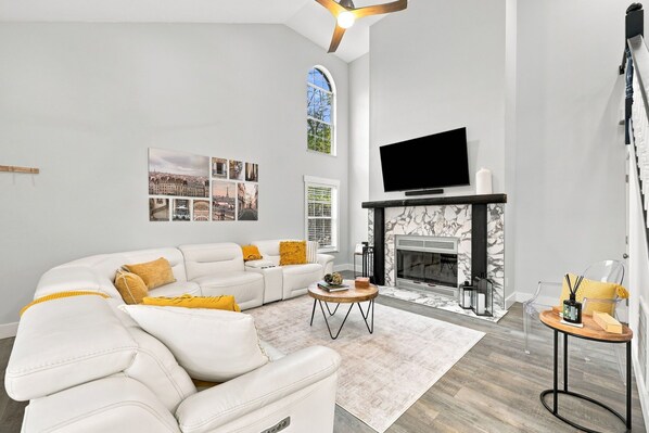 A modern living room with a sectional sofa, a fireplace, a flat-screen TV, and a large window with a cityscape view.