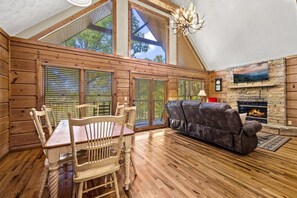 Cozy cabin-style interior with a high ceiling and large windows, featuring a dining table, sofa, and stone fireplace.