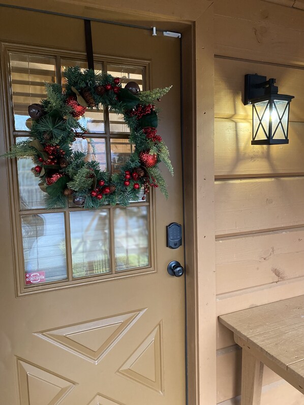 A beige door with a Christmas wreath, a security system sign, and a wall-mounted lantern. - Family Friendly Cabin HotTub Games, Minutes to Dollywood & The Parkway. (Sevierville)