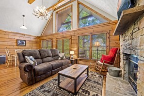 Cozy living room with a high ceiling, large windows, and a fireplace, featuring a leather sofa, armchair, and wooden coffee table.