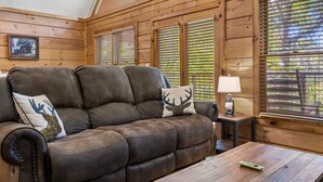 Ein gemütliches Holzhaus-Interieur mit einem großen braunen Ledersofa, zwei Zierkissen, einem kleinen Tisch mit Lampe und einem gerahmten Bild an der Wand.