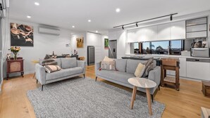 A modern living room with a grey sofa, a small round table, and a kitchen with a large window. - StayCentral - East Brunswick Penthouse with Study (Brunswick East)