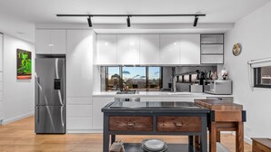 A modern kitchen with a stainless steel refrigerator, a large island with a sink, and a view of the city through large windows. - StayCentral - East Brunswick Penthouse with Study (Brunswick East)