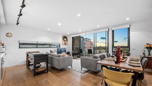 A modern living room with a dining area, large windows, and a view of the cityscape. - StayCentral - East Brunswick Penthouse with Study (Brunswick East)