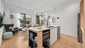 A modern kitchen with a built-in oven, a dining area with a sofa and coffee table, and a living room with a TV and a plant. - StayCentral - Boulevard Gardens (Coburg)