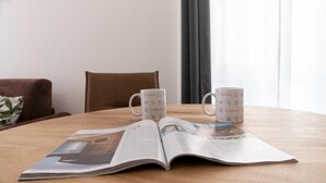 Dos tazas blancas con la marca Italianway sobre una mesa de madera, una revista abierta con texto e imágenes, y una silla marrón al fondo.
