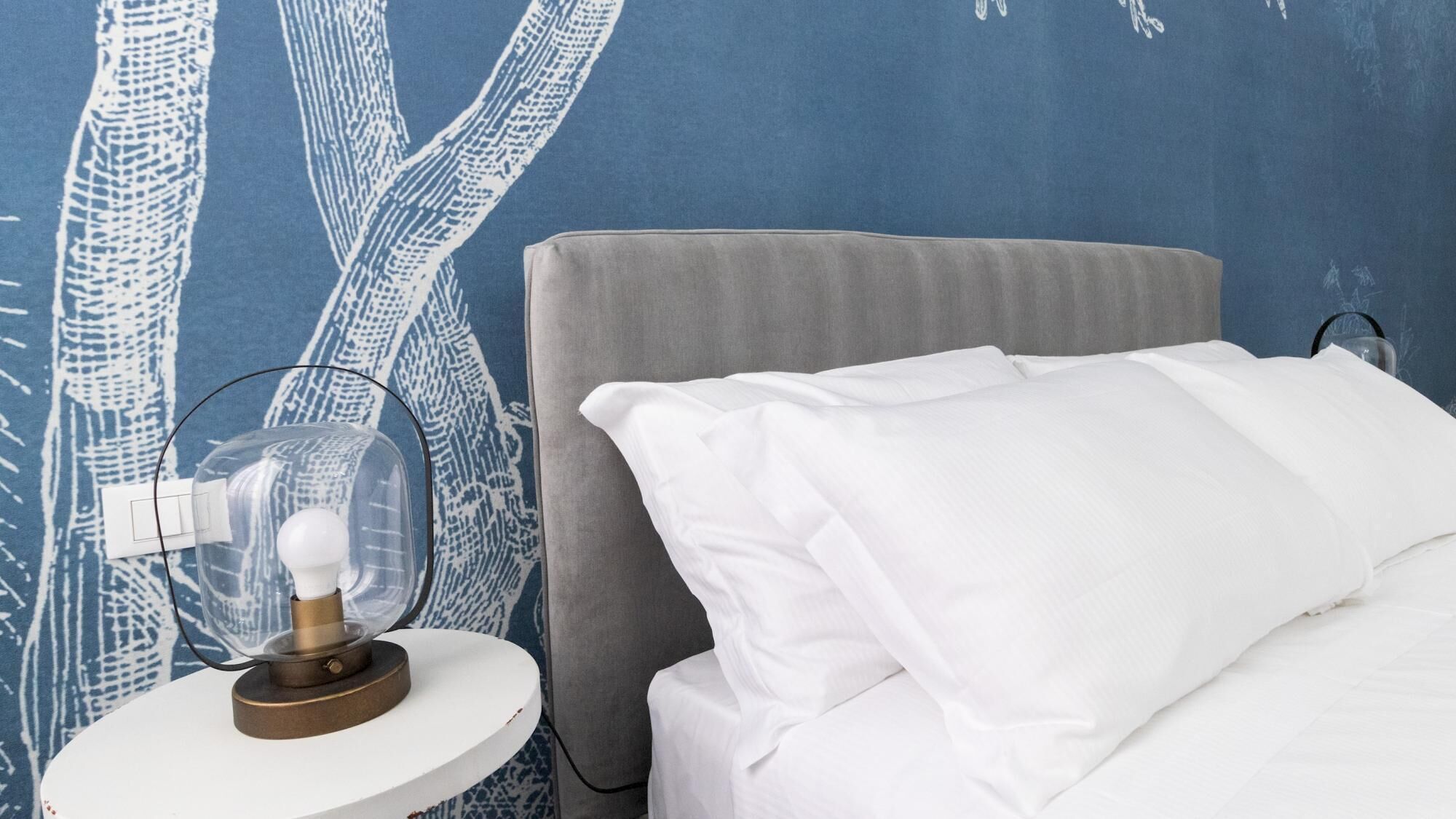 Una cama bien tendida con sábanas blancas, una mesita de noche redonda con una lámpara, y una pared con un diseño abstracto azul.