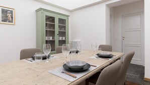 Una mesa de comedor dispuesta con platos, tazones y copas de vino, rodeada de sillas. En el fondo, un aparador verde con puertas de cristal.