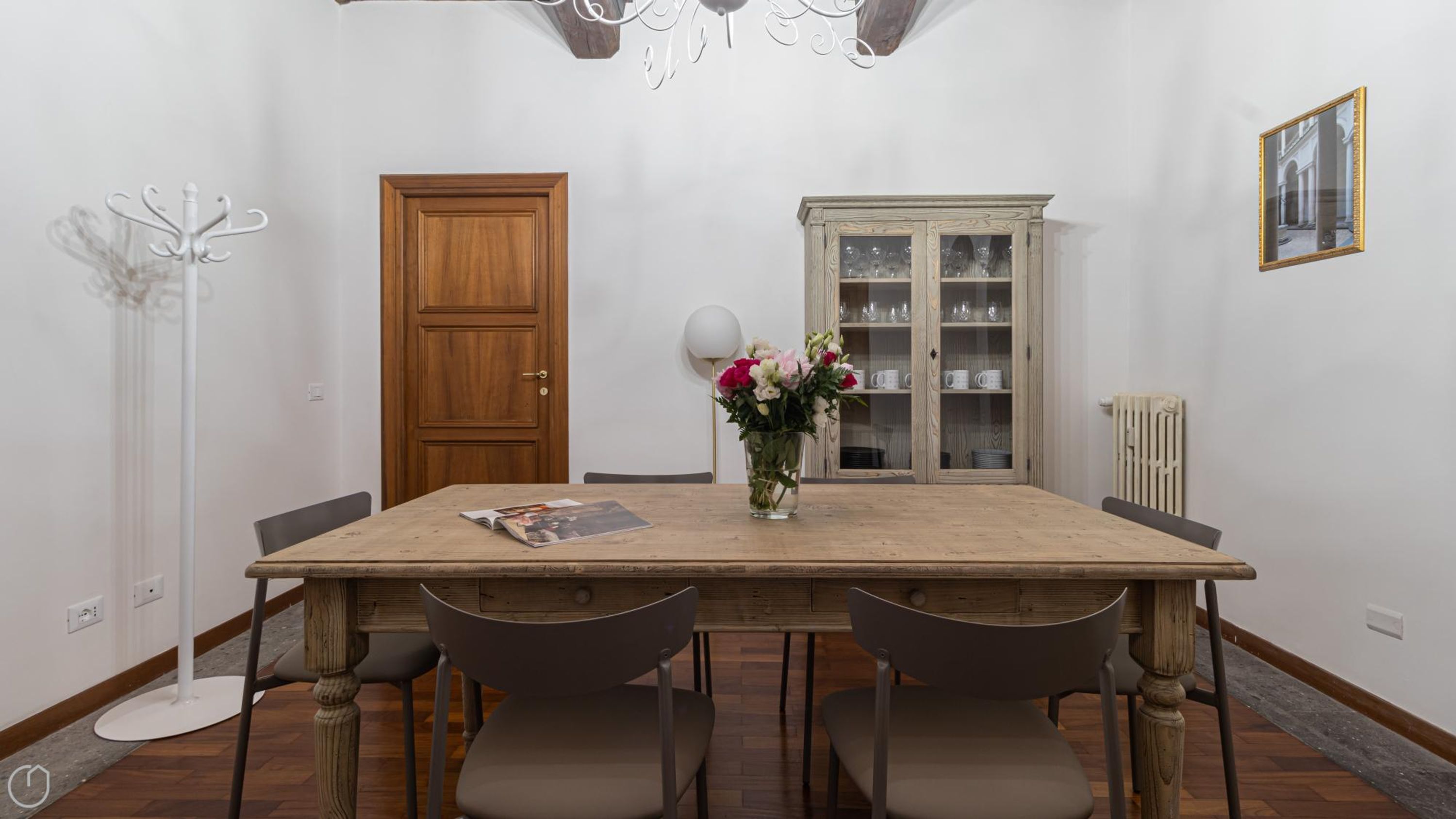 Ein Esszimmer mit Holztisch, vier Stühlen, einer Blumenvase, einem Schrank mit Glastüren, einem Garderobenhaken und einem gerahmten Bild an der Wand.