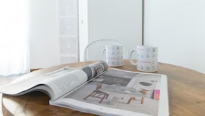 A table with a newspaper, two white mugs with Italianway logos, and a white chair.