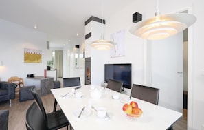 A modern dining area with a white table set for four, a fruit bowl, and a large flat-screen TV on the wall. - Promenadenvilla (Lübeck)