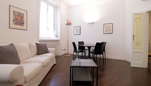 A modern living room with a white sofa, a glass-top coffee table, and a dining area with a small table and four chairs. - Italianway Easy - Vanvitelli 49 (Milan)