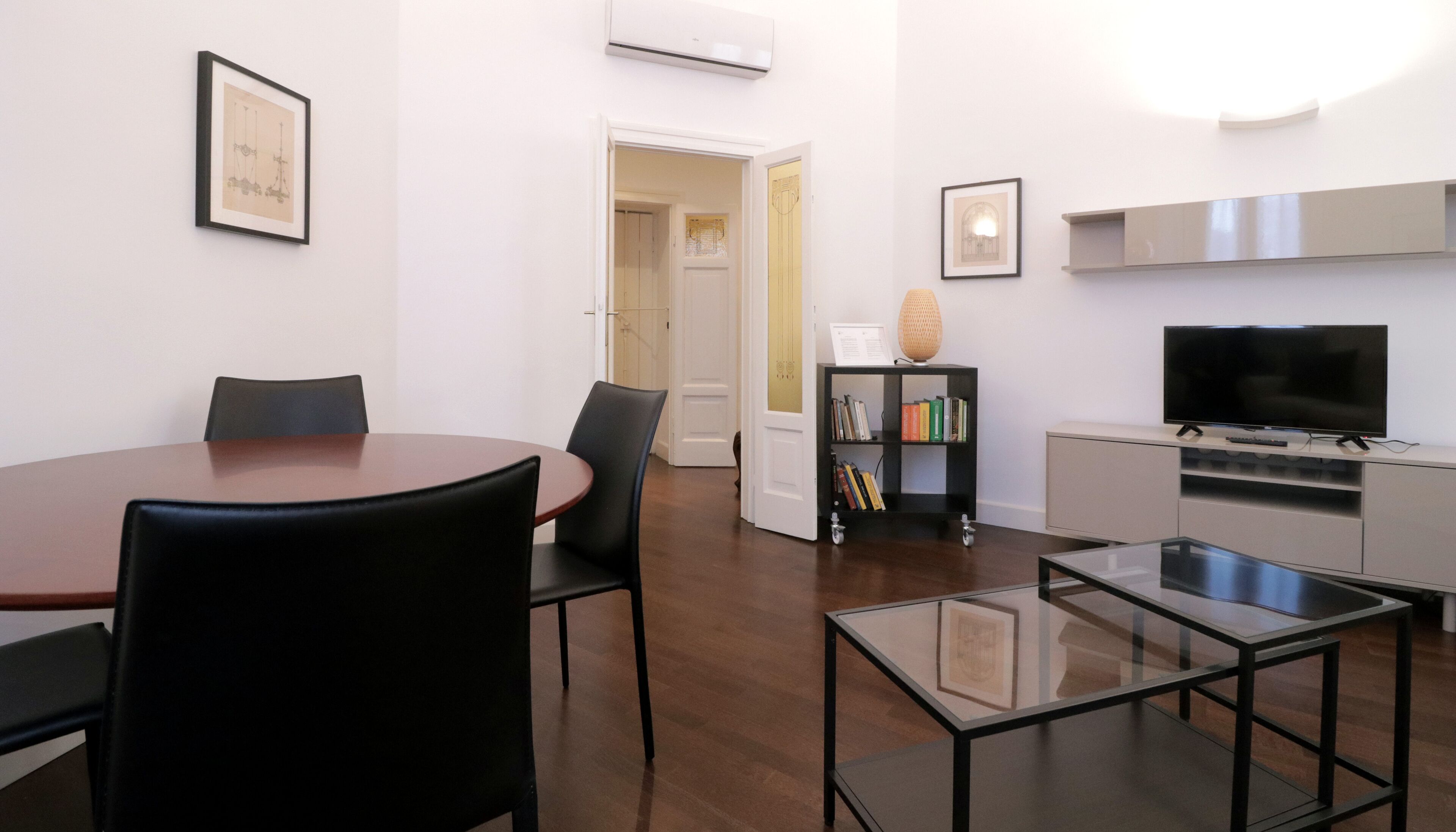 Un salon moderne comprenant une table à manger, des chaises, une table basse en verre, une bibliothèque, un téléviseur et un coin cuisine.