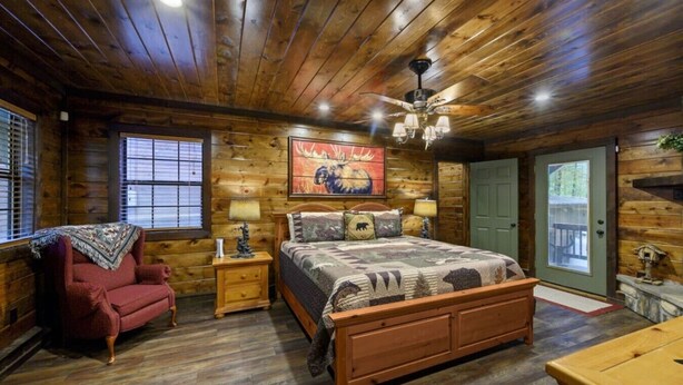 A wooden cabin bedroom with a bed, nightstands, a chair, and a door. - Bent Pine Lodge | Spacious 4BR Broken Bow Cabin with Hot Tub, Fire Pit & Grill (Hochatown)