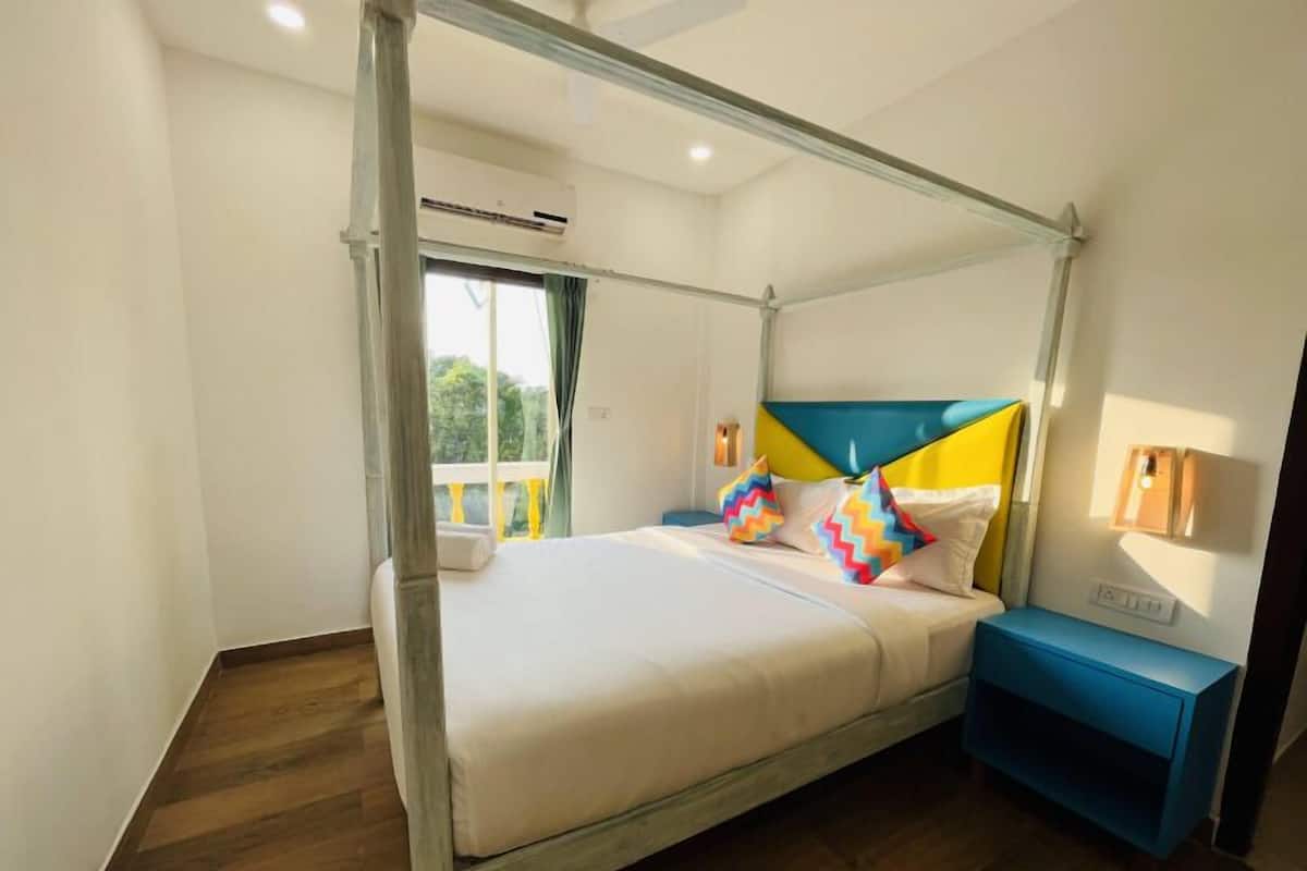 A four-poster bed with a white mattress and colorful pillows, a blue nightstand, and a window with curtains.