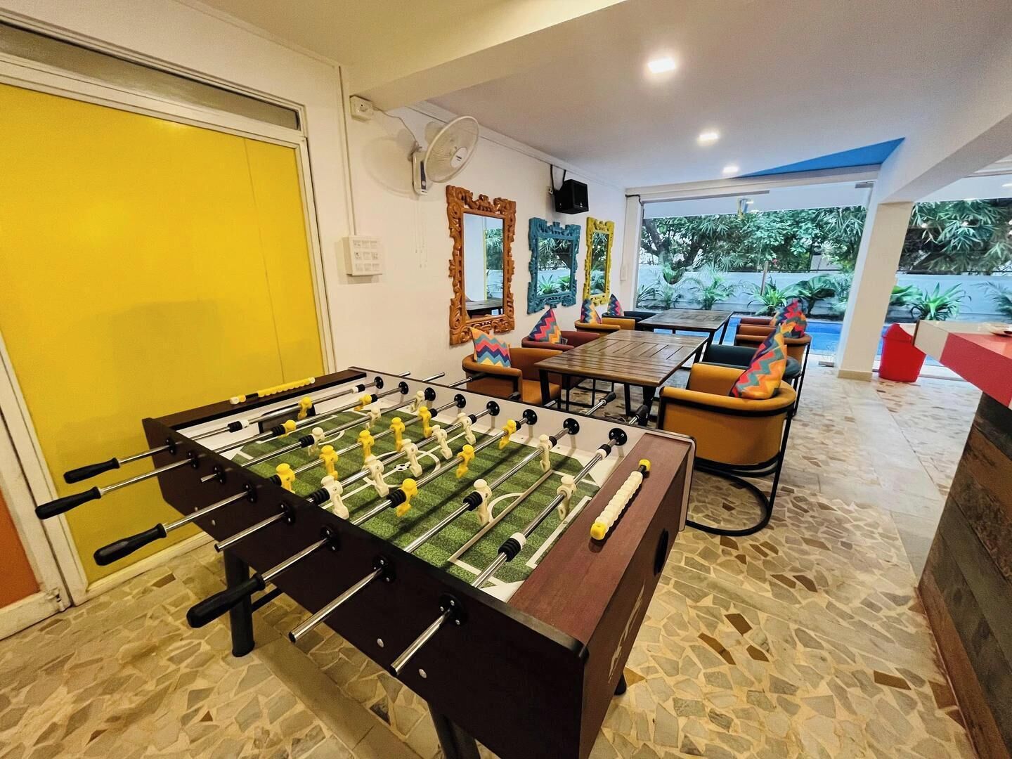 Una mesa de fútbol en una habitación con una puerta amarilla, piso de baldosas de piedra y vista a zonas verdes.
