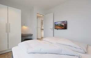 A bedroom with a bed, a wardrobe, a desk, and a wall-mounted TV. - Dünenvilla (Lübeck)