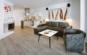 A modern living room with a grey sofa, a coffee table, and a grey armchair. There is a kitchen area with white cabinets and a stainless steel range hood. - Dünenvilla (Lübeck)