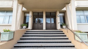 Ein Gebäudeeingang mit der Aufschrift RHODORA über der Tür, eine Treppe, die zum Eingang führt, und je eine Pflanzengruppe auf beiden Seiten.