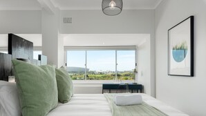 Ein modernes Schlafzimmer mit einem großen Bett, grünen Kissen, Blick auf eine Stadtlandschaft durch ein großes Fenster und einem gerahmten Bild an der Wand.