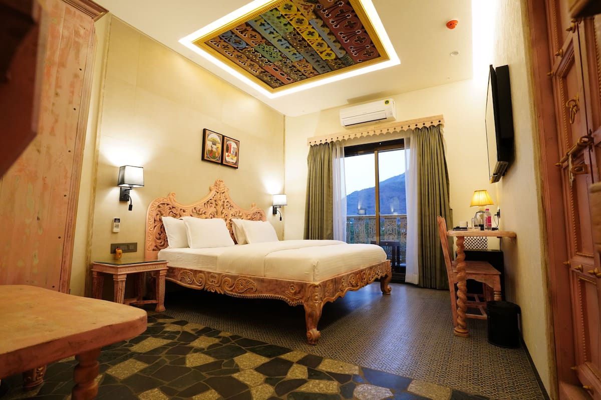 A hotel room with a large bed, a wooden desk, a chair, a small table, a nightstand, a wall-mounted TV, and a window with curtains.