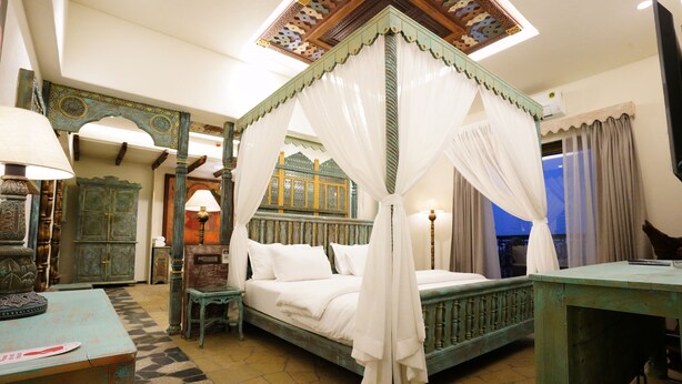 A four-poster canopy bed with white linens, a wooden nightstand, and a small table with a lamp. - The Kinara Village (Mawal)