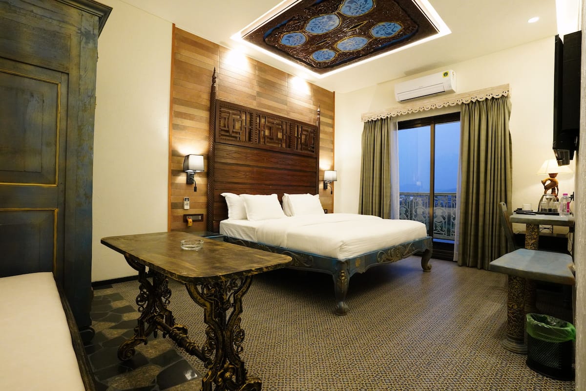 A hotel room with a large bed, a wooden headboard, a small table, a cat tree, and a view of the city through the window.