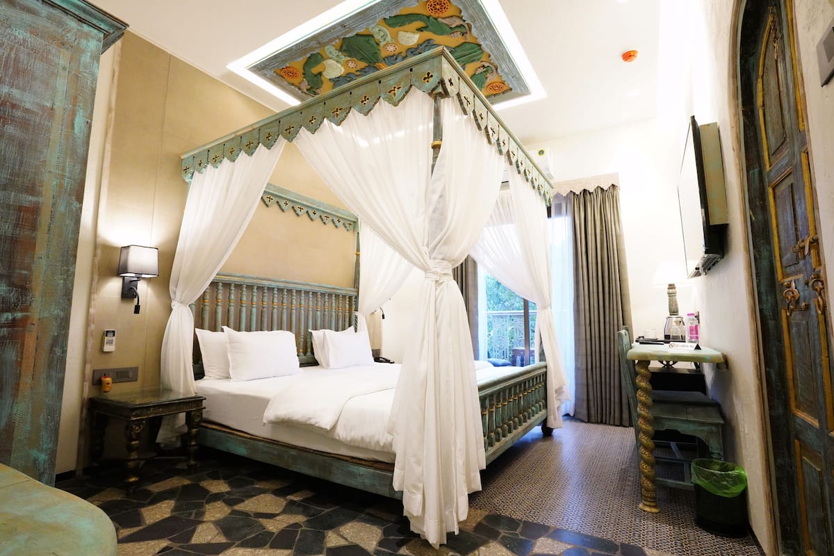 A four-poster bed with white linens, a nightstand, a small table with a lamp, and a green trash bin.