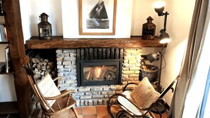 Una chimenea acogedora con un marco de piedra, dos sillas de madera con cojines, un cuadro enmarcado sobre la chimenea y un farol en la repisa.