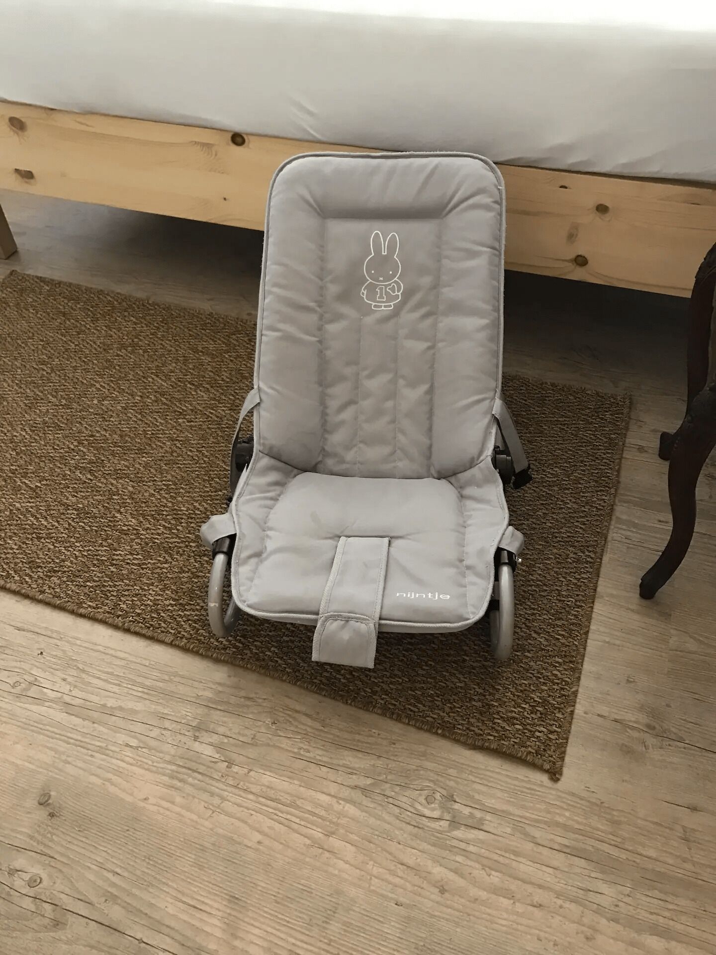 Un transat pour bébé de couleur grise, orné d’un motif de lapin, est placé sur un tapis marron dans une pièce dont le sol est en bois, avec un lit visible au fond.