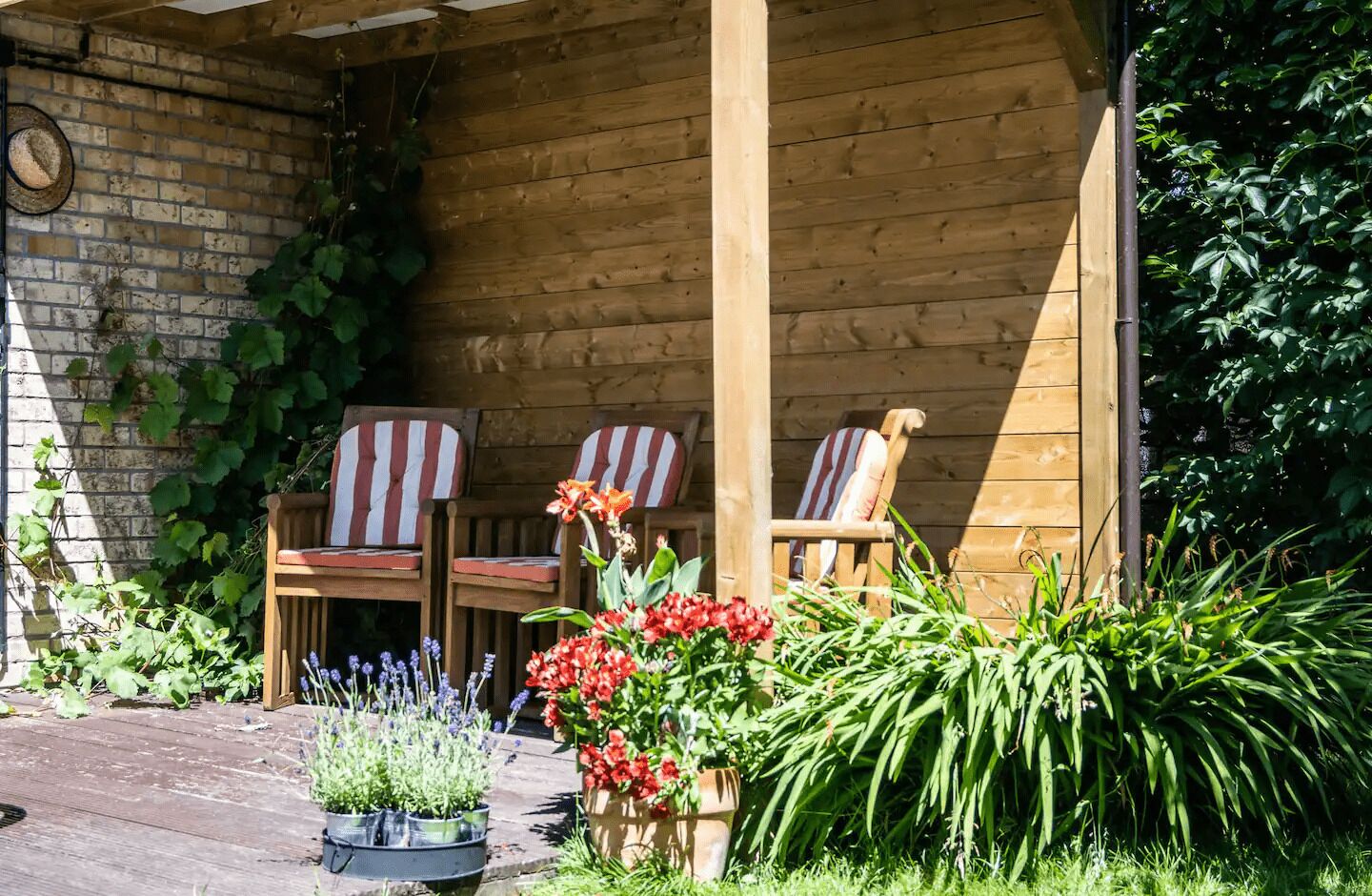 A wooden porch with three striped chairs, potted plants, and a brick wall.