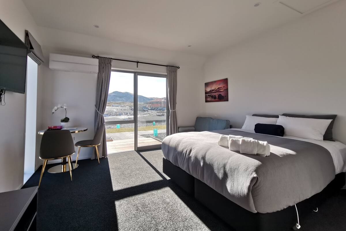 A modern hotel room with a large bed, a desk, a chair, and a view of the outdoors.