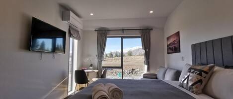 A modern hotel room with a large bed, a flat-screen TV, a desk, and a view of a mountainous landscape through the window.