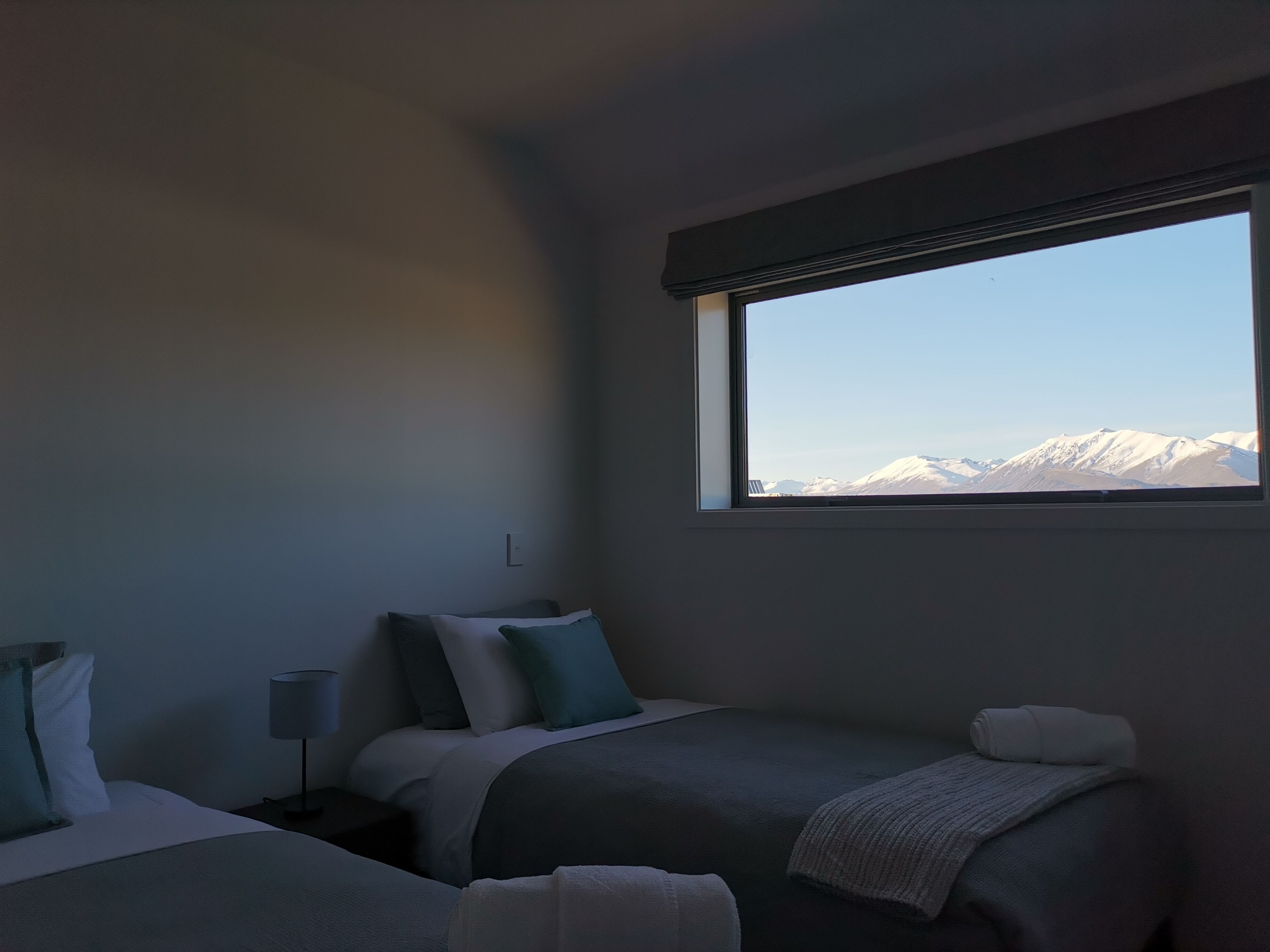 Quarto de hotel com duas camas, uma janela com vista para as montanhas e uma mesa de cabeceira com abajur.