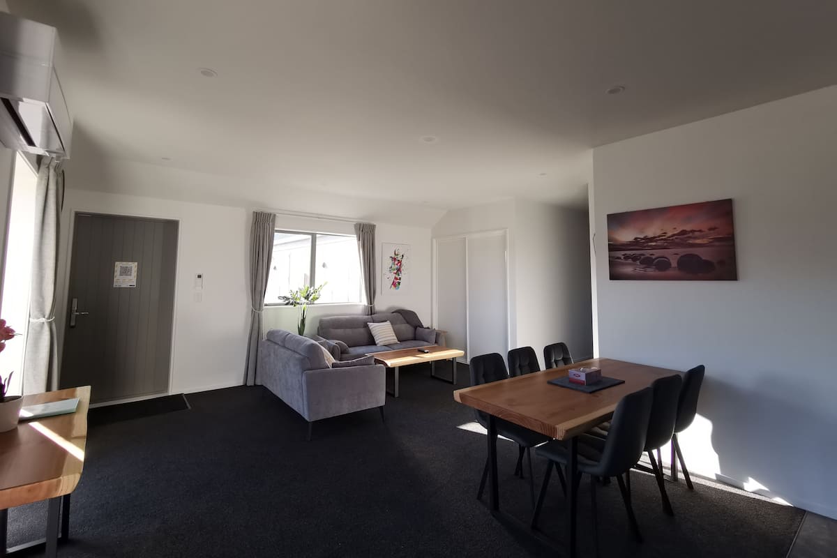 A modern living room with a grey sofa, a wooden dining table with chairs, a small coffee table, and a painting on the wall.