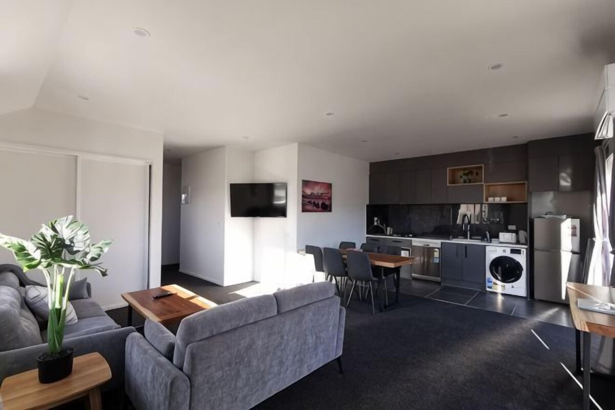 A modern living room with a sofa, coffee table, and a TV. It includes a kitchen with a dining area and a washing machine.