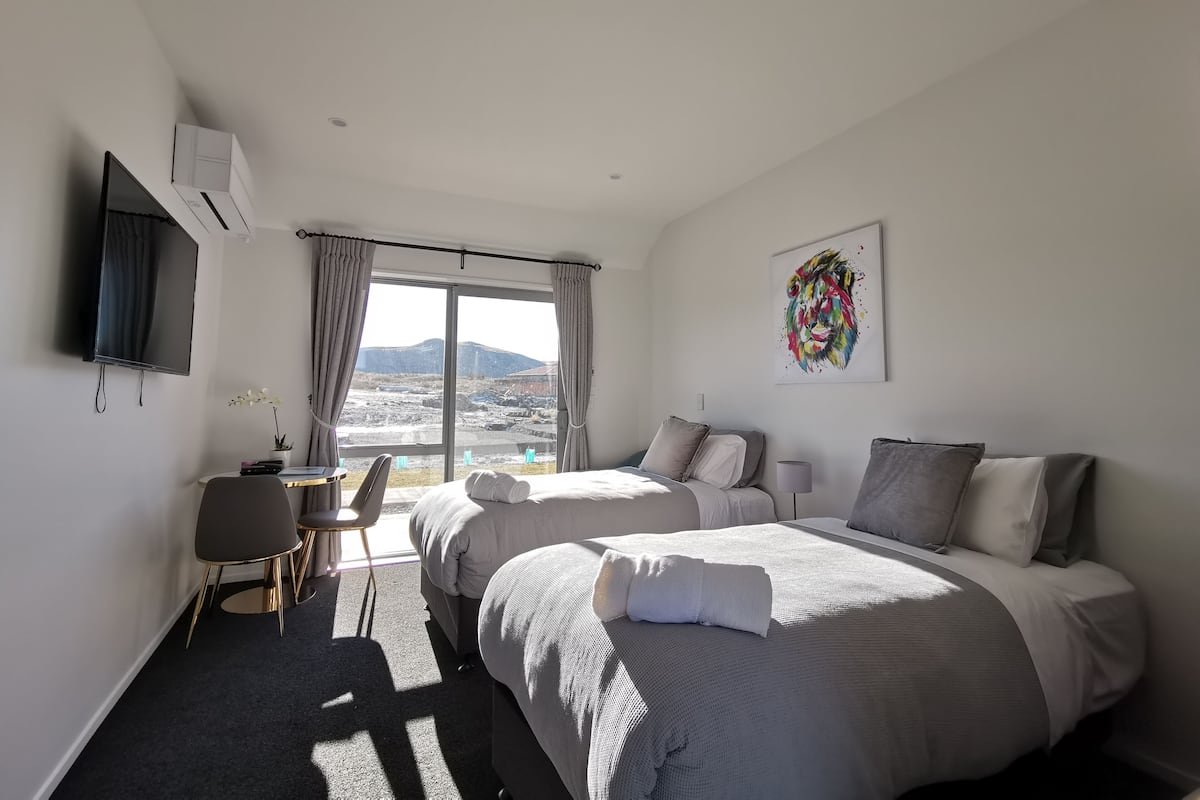 A hotel room with two beds, a desk, a chair, a TV, and a large window with a mountain view.