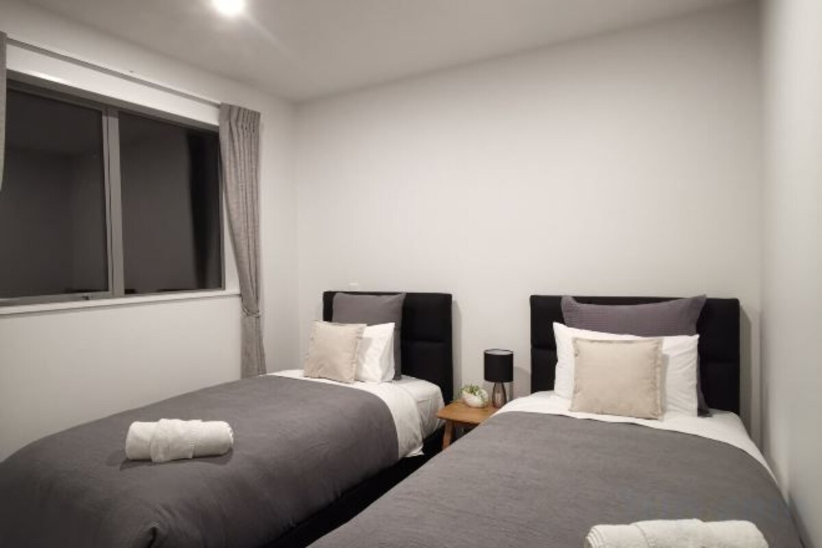 A hotel room with two single beds, a window with curtains, a bedside table with a lamp, and a small nightstand.