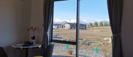 A room with a bed, a small table with a vase, and a view of mountains through large windows.