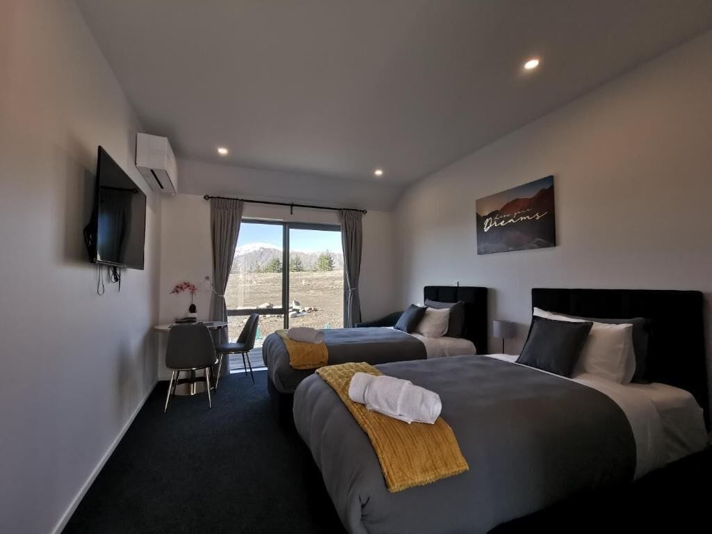 A hotel room with two beds, a desk, and a chair. There is a window with a view of the outdoors.