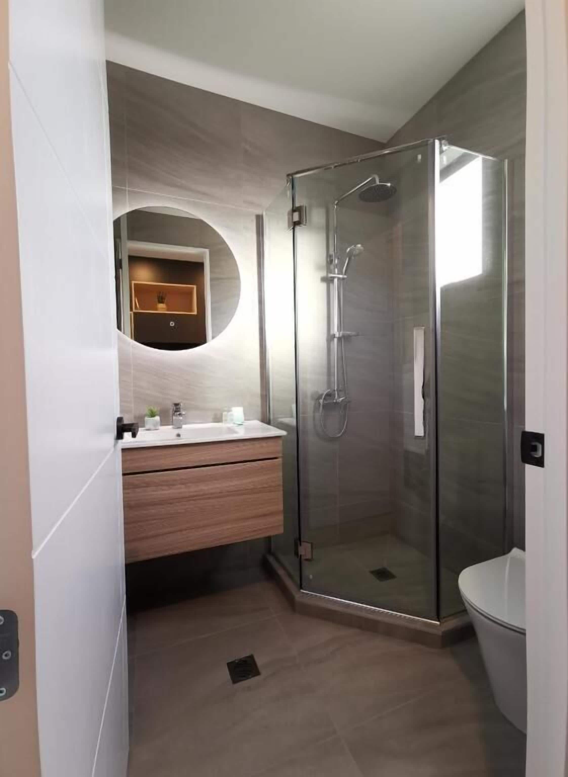 A modern bathroom with a glass-enclosed shower, a round mirror, and a wooden vanity.