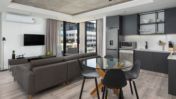 A modern living room with a sectional sofa, a glass dining table, and a kitchen area with a microwave and dishwasher. - The Signature 414 (Cape Town)