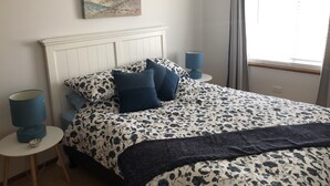 Et soveværelse med seng, et natbord, en lampe, et billede på væggen og et vindue med gardiner.
