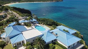 Une propriété située sur la côte, comprenant plusieurs bâtiments aux toits bleus, une piscine et offrant une vue sur l’océan et la plage.