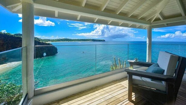 Un espace extérieur abrité doté d’une terrasse en bois, d’un fauteuil confortable et d’une balustrade en verre, offrant une vue sur une mer d’un bleu profond et une côte rocheuse.