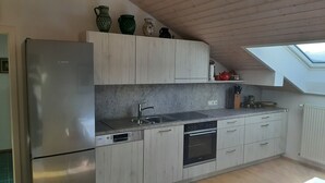 A modern kitchen with a stainless steel refrigerator, a built-in dishwasher, and an oven. There is a skylight above the sink area, and wooden cabinets with a light finish. - Apartment with 1 sep. Bedroom, southwest-facing roof terrace, 58 sqm (Prien am Chiemsee)