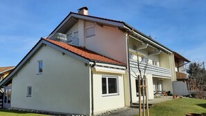 A two-story house with a red-tiled roof, a balcony, and a small front porch. - Apartment with 1 sep. Bedroom, southwest-facing roof terrace, 58 sqm (Prien am Chiemsee)